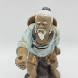 May include: A ceramic figurine of an elderly person with a white beard and hair, wearing a light blue tunic and brown patterned shorts. The figure has a tan complexion, a dark topknot, and is posed with one arm extended forward with a small hole.