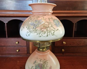 May include: Antique oil lamp with a pink and white glass shade. The shade features a floral design in shades of blue, green, and purple. The lamp has a brass base and a clear glass chimney. A vintage decorative piece.