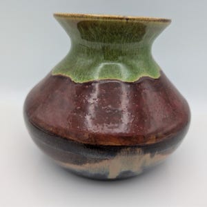 May include: A small ceramic vase with a wide, rounded body and a flared rim. The glaze features a layered color scheme with a moss green top section, a deep reddish-brown middle, and a dark brown base with subtle cream-colored drip accents.