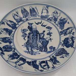 May include: A white ceramic plate with a blue painted design. The plate features a central image of a bearded figure with other figures around the rim. The plate has a blue border.