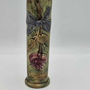 May include: A cylindrical vase with a floral design. The vase is primarily green with a dark blue bow and a maroon flower. The vase has a textured surface and a rounded base. The vase is approximately 8 inches tall.