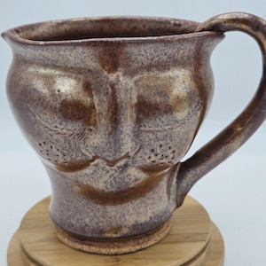 May include: A ceramic mug with a speckled brown and tan glaze featuring a sculpted face on the front. The mug has a wide rim, a curved handle on the right side, and a rounded body. The face includes embossed eyes, a nose, and dotted cheeks.