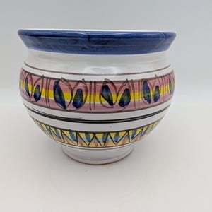 May include: A white ceramic planter with a rounded body and a flared rim. The exterior features a dark blue rim, horizontal pink and yellow stripes, and hand-painted dark blue leaf motifs arranged in a repeating pattern around the center.