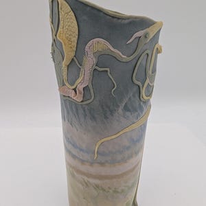 May include: A tall, ceramic vase with a unique design. The vase features a muted color palette of blues, greens, and beige, with raised, textured sea creature-like embellishments in shades of yellow and pink. The vase has a slightly angled top.