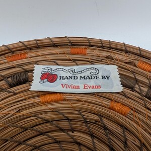 May include: Close-up of a handmade woven basket with a label that reads "HAND MADE BY Vivian Evans." The basket is made of brown and tan natural fibers, with orange and brown stitching accents. The label features a red yarn ball.
