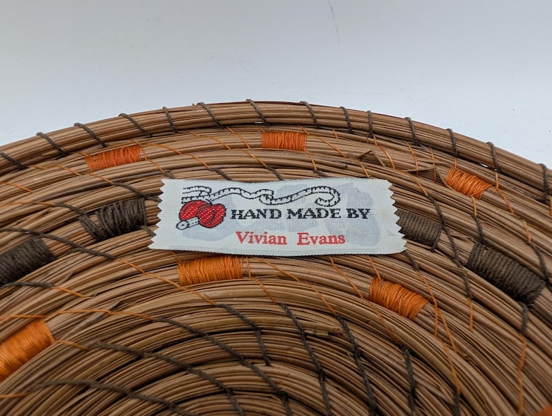 May include: Close-up of a handmade woven basket with a label that reads "HAND MADE BY Vivian Evans." The basket is made of brown and tan natural fibers, with orange and brown stitching accents. The label features a red yarn ball.