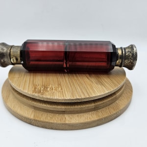May include: A rectangular, ruby-red glass perfume bottle with ornate, silver-toned metal caps on each end. The bottle rests on a three-tiered, round bamboo display stand. The glass has a faceted design.