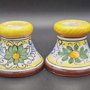 May include: A pair of ceramic candle holders featuring a flared base and a rounded top rim. The surface is decorated with hand-painted floral motifs in green, yellow, and blue against a white background, finished with a solid brown base trim.