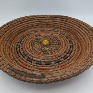 May include: A round, woven basket with a spiral design. The basket is made of brown material with accents of black, orange, and a yellow center. The basket is likely handmade and suitable for decorative or functional use.
