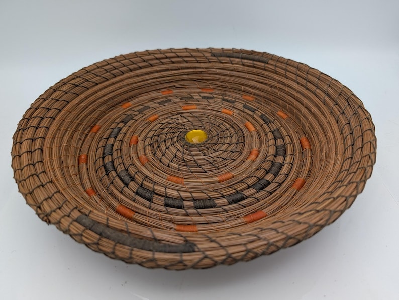 May include: A round, woven basket with a spiral design. The basket is made of brown material with accents of black, orange, and a yellow center. The basket is likely handmade and suitable for decorative or functional use.