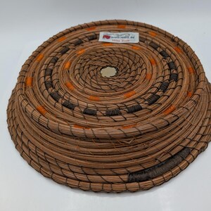 May include: A round, woven basket with a spiral design. The basket is made of natural brown fibers, with accents of orange and dark brown. The basket has a label that reads "Hand crafted by Vera Davis."