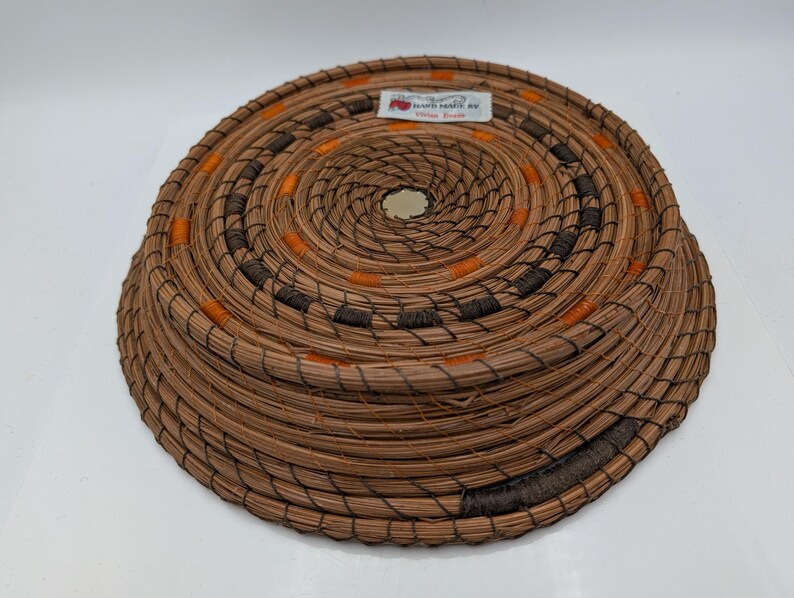 May include: A round, woven basket with a spiral design. The basket is made of natural brown fibers, with accents of orange and dark brown. The basket has a label that reads "Hand crafted by Vera Davis."