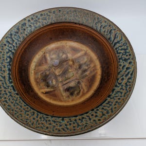 May include: A round ceramic bowl featuring a wide, textured rim with a mottled blue and brown pattern. The interior of the bowl has a smooth, deep brown glazed finish with a central cross-shaped indentation and subtle variations in color tones.