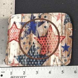 Puede incluir: Una cartera rectangular con un patrón de estrellas patrióticas en rojo, blanco y azul. La cartera tiene un diseño circular marrón en el centro. La cartera mide aproximadamente 14 cm de ancho.