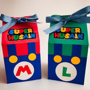 Może przedstawiać: Dwa pudełka na prezenty w stylu Super Mario. Jedno jest czerwono-niebieskie z dużym białym okręgiem i czerwoną literą "M". Drugie jest zielono-niebieskie z białym okręgiem i zieloną literą "L". Oba mają napis "SUPER HUSAIN" i są zawiązane niebieskimi wstążkami.
