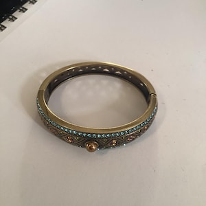 May include: A gold-toned hinged bangle bracelet featuring a central row of light blue crystals, a geometric lattice pattern with embedded multi-colored crystals, and a single smooth gold-toned sphere accent on the front surface.