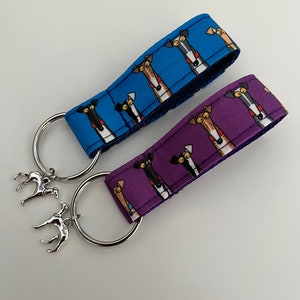 May include: Two keychains with silver rings and silver dog charms. One keychain has a blue strap with a repeating pattern of cartoon dogs. The other keychain has a purple strap with a repeating pattern of cartoon dogs.