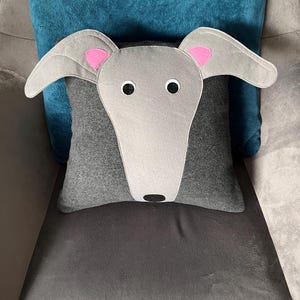 May include: A grey felt cushion with a grey felt dog face on it. The dog has pink ears and black eyes. The cushion is sitting on a grey sofa.