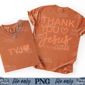 May include: Two rust-colored t-shirts with the text "THANK YOU JESUS" and "TY" printed on them. A silver chain necklace with a toggle clasp is draped over one shirt. The shirts are on a white background.