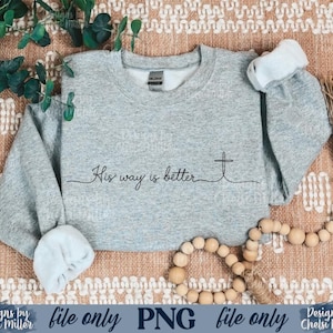 May include: A light gray sweatshirt with the text "His way is better" and a cross design. The long-sleeved crew neck top has rolled-up cuffs. The image also includes a string of wooden beads and greenery.