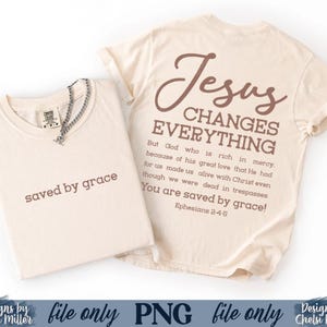 K&ouml;nnte beinhalten: Zwei cremefarbene T-Shirts. Eines ist gefaltet, mit "saved by grace" in Braun gedruckt. Das andere Shirt hat "Jesus CHANGES EVERYTHING" und einen Bibelvers auf der R&uuml;ckseite. Eine silberne Halskette befindet sich auf dem gefalteten Shirt.