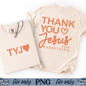 May include: Two cream-colored t-shirts with orange text. One shirt reads "TYJ" with a heart, and the other says "THANK YOU Jesus FOR EVERYTHING." A silver necklace with a pendant is on the first shirt. The shirts are folded.