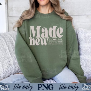 Könnte beinhalten: Ein olivgrünes Sweatshirt mit dem Schriftzug "Made new" in einer großen, stilisierten Schriftart. Darunter befindet sich der Bibelvers "2 COR 5:17". Das Sweatshirt ist einfarbig und hat lange Ärmel.