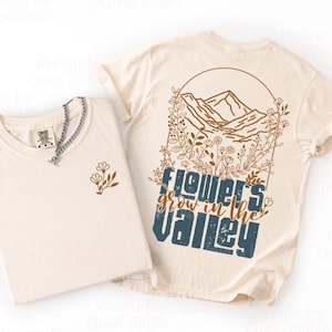 Könnte beinhalten: Zwei cremefarbene T-Shirts. Ein Shirt hat ein kleines Blumendesign auf der Vorderseite und eine silberne Halskette. Das andere Shirt hat ein Berg- und Blumendesign mit den Worten "Flowers grow in the Valley" in Blau und Braun.