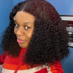 May include: A woman wearing a curly black wig with a lace front. The wig has a natural-looking part and is styled with voluminous curls. She is wearing a red and white striped jacket.
