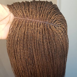 May include: A brown braided wig with a lace front. The wig features numerous small, tightly woven braids, creating a textured appearance. The color is a mix of light and dark brown tones.