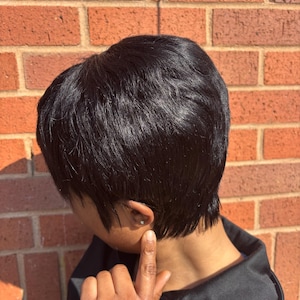 Puede incluir: Una persona con el pelo corto y negro peinado en un corte pixie. El pelo es liso y recto, con una ligera raya lateral. La persona lleva una chaqueta negra con rayas blancas en las mangas.