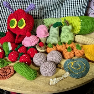 May include: A collection of colorful, crocheted food items and a red caterpillar toy. The food items include strawberries, cupcakes, pears, pumpkins, and a slice of cheese. The caterpillar has a red body, green eyes, and purple antennae.