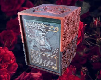 May include: A red and black wooden box with a decorative rose pattern. The box has a clear plastic window that shows a Magic: The Gathering card with the text "Mana Apex of Wishes".
