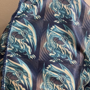 May include: A fabric item featuring a repeating pattern of a blue and white dragon design. The dragon has a fierce expression and is set against a dark blue background. The edges are trimmed with a darker blue.
