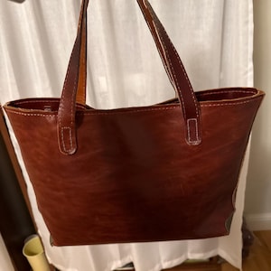 May include: A brown leather tote bag with two handles. The bag has a rectangular shape with a slightly wider opening. The handles are attached with white stitching. The leather has a textured appearance.