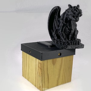May include: A black 3D printed gargoyle statue sits atop a wooden box with a black lid. The box has a solar panel on top and is designed for outdoor use.