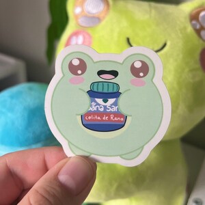 May include: A green frog sticker holding a blue bottle with the text "Sana San" and "coliita de Rana".