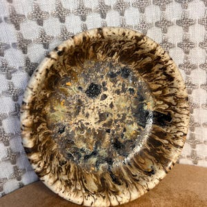 Peut inclure: Un petit plat rond en céramique avec un design marbré unique. Le plat présente un mélange de couleurs marron, noir et crème, créant un motif tourbillonnant. Le bord est de couleur crème plus claire. Ce plat décoratif est adapté pour contenir de petits objets.