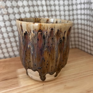 May include: A handmade ceramic cup with a unique glaze. The cup has a beige rim that drips down into shades of brown, black, and purple. The bottom of the cup is a creamy white colour. The cup is on a wooden surface.