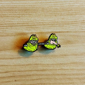 Può includere: Una spilla in smalto verde neon raffigurante due fantasmi in stile cartoon collegati da una barra di metallo. I fantasmi hanno denti bianchi, contorni neri e uno tira fuori la lingua. La spilla è posta su uno sfondo di legno.