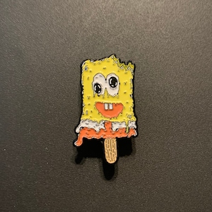 May include: A yellow enamel pin shaped like a melting popsicle featuring a cartoon character with large eyes and an orange smile. The pin has a wooden stick and is set against a dark background.