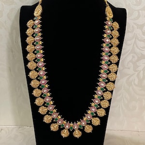 May include: A gold-colored necklace and matching earrings set. The necklace features coin-shaped pendants with intricate designs, interspersed with red and green gemstone accents. The earrings have a similar design. The jewelry is displayed on a black mannequin.