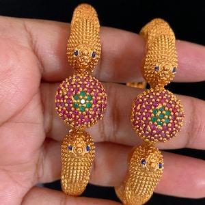 May include: Two gold-toned bangles featuring textured animal head motifs at the ends. Each bangle is centered with a circular medallion encrusted with small pink and green stones. The animal heads have tiny blue stone eyes and a detailed scale pattern.