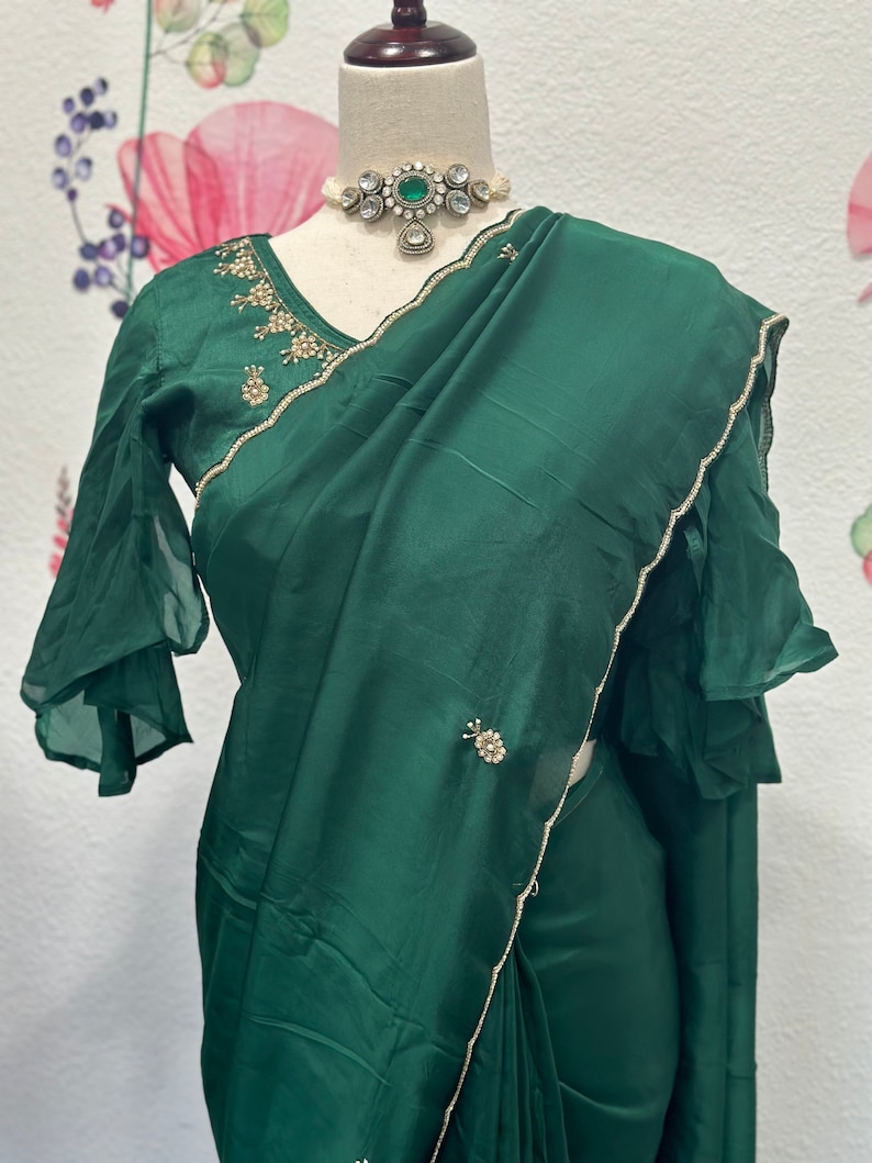 May include: A dark green silk saree with a gold embroidered border and embellishments. The saree has a ruffled sleeve blouse with a matching gold embroidered border.