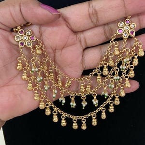 May include: A gold-toned multi-strand necklace featuring two floral-shaped end pieces set with pink and clear stones. The three cascading chains are adorned with numerous small gold-toned bell-shaped charms and tiny white and green bead accents.