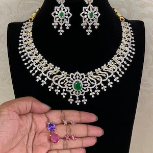May include: A jewelry set featuring a necklace and matching earrings with silver-toned metal, clear crystals, and a central oval green stone. Additional loose oval stones in purple and pink are displayed alongside the main jewelry set.