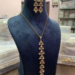 May include: A gold-toned jewelry set displayed on a blue velvet bust. The set includes a long pendant necklace and matching earrings. The necklace and earrings feature a cascading design with small white pearls and pink accents.