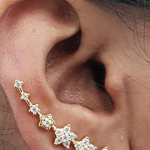 May include: A gold-colored ear climber earring featuring a series of small, sparkling clear gemstones arranged in a floral and starburst design. The earring is worn on the ear, showcasing its delicate and intricate details. The jewelry is set against the backdrop of the ear.