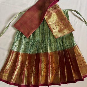 May include: A pleated skirt featuring a green upper section with a subtle floral pattern and a wide, deep red lower border adorned with intricate gold metallic woven designs. A matching red and gold fabric piece is draped across the top.