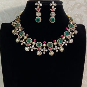 May include: A jewelry set consisting of a necklace and matching drop earrings. The pieces feature round and square-cut green and pink stones set in a metallic frame, accented with small clear stones arranged in a floral-inspired pattern.